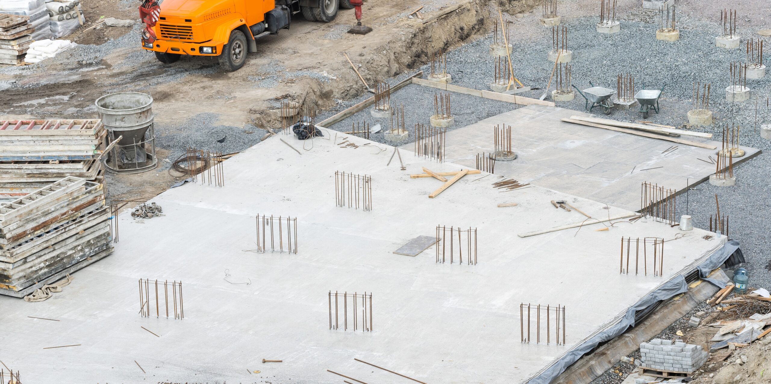 Concreting,Work:,Construction,Site,Worker,During,Concrete,Pouring,Into,Formwork A construction site with concrete foundation, rebar protruding, materials stacked, and an orange truck in the background.